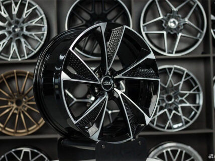 Close-up of a glossy black alloy wheel with a bold multi-spoke design and Audi center cap, on display with other rims in the background.