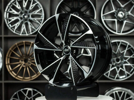 Glossy black alloy wheel with angular spokes and Audi center cap on display, surrounded by other rims on a showroom wallide.
