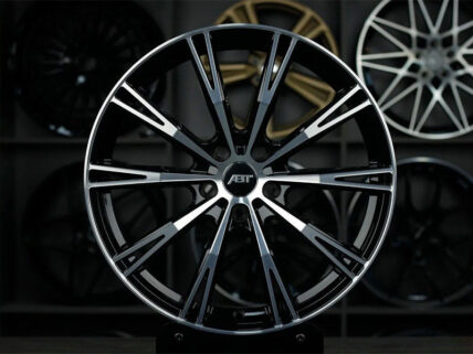 Close-up of a large black and silver multi-spoke alloy car wheel with an ABT center cap in a showroom backdrop of other rims.