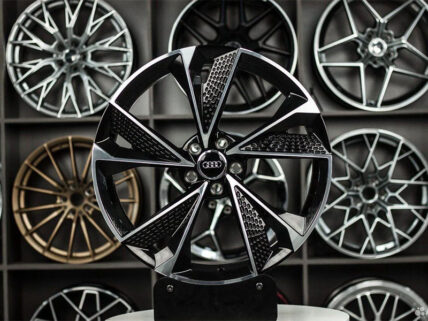 Close-up of a black and silver Audi alloy wheel on a display stand, with a wall of other rims in the background.
