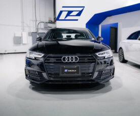 Front view of a black Audi sedan in a bright showroom, Tunezilla plate, blue logo on wall behind.