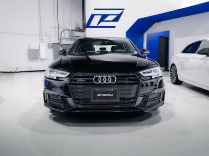Front view of a black Audi sedan in a bright showroom, Tunezilla plate, blue logo on wall behind.